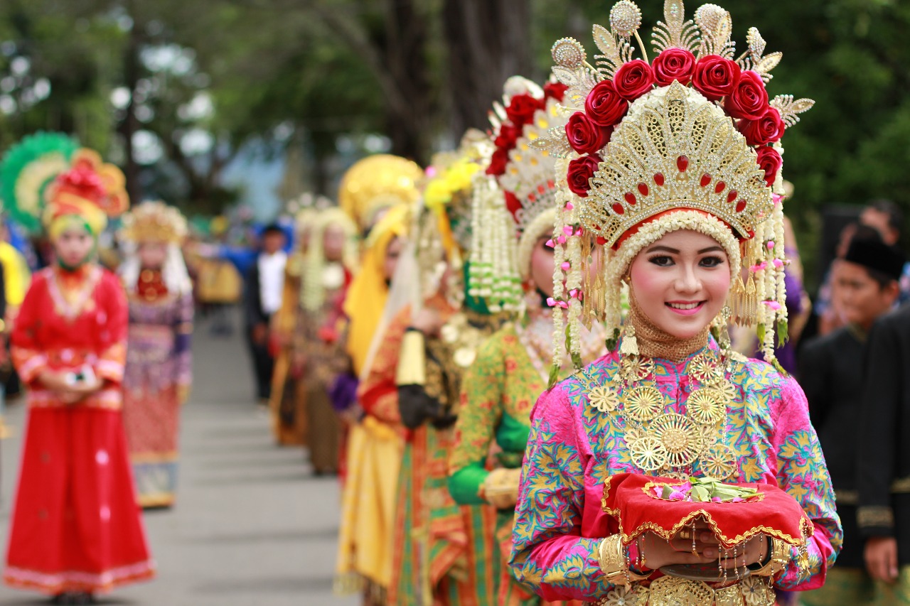  A smiling woman wearing a traditional Indonesian headdress and clothing, surrounded by other people in traditional dress, during a parade.
